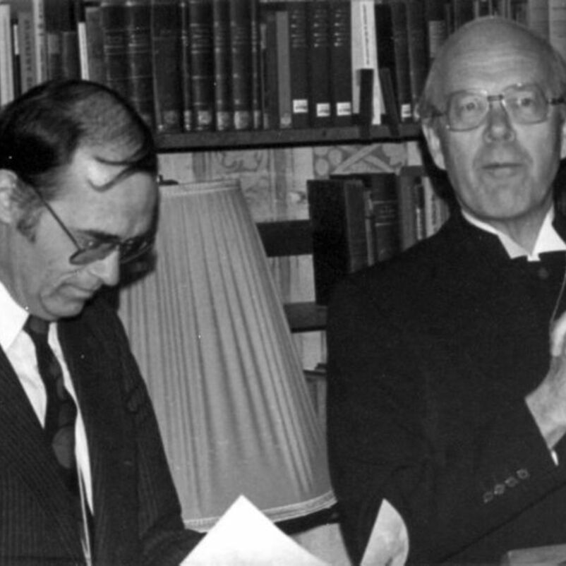 Das Schwarzweiß-Bild zeigt zwei männlich lesbare Personen in schwarzen Anzügen sitzend vor einer dicht gefüllten Bücherwand.