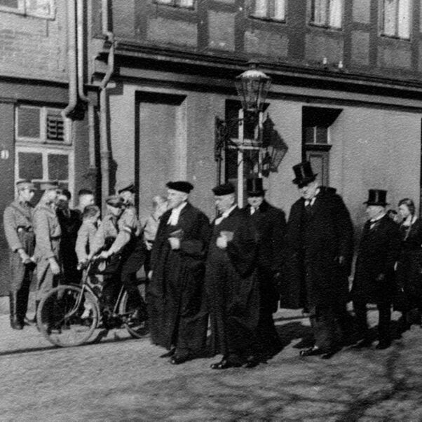 Das Schwarzweiß-Bild zeigt eine Gruppe von männlich lesbaren Pastoren im Talar, die auf der Straße eine Gruppe von Jugendlichen in HJ-Uniformen begegnen.