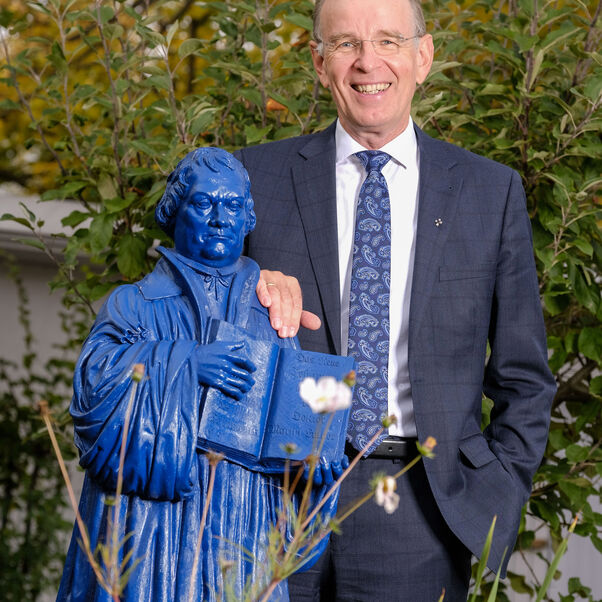 Das Bild zeigt eine männlich lesbare Person im Garten stehend und an einer blauen Luther-Statue lehnend. Die Person lächelt und trägt einen dunklen Anzug und ein helles Hemd, hat helle kurze Haare und eine Brille. 