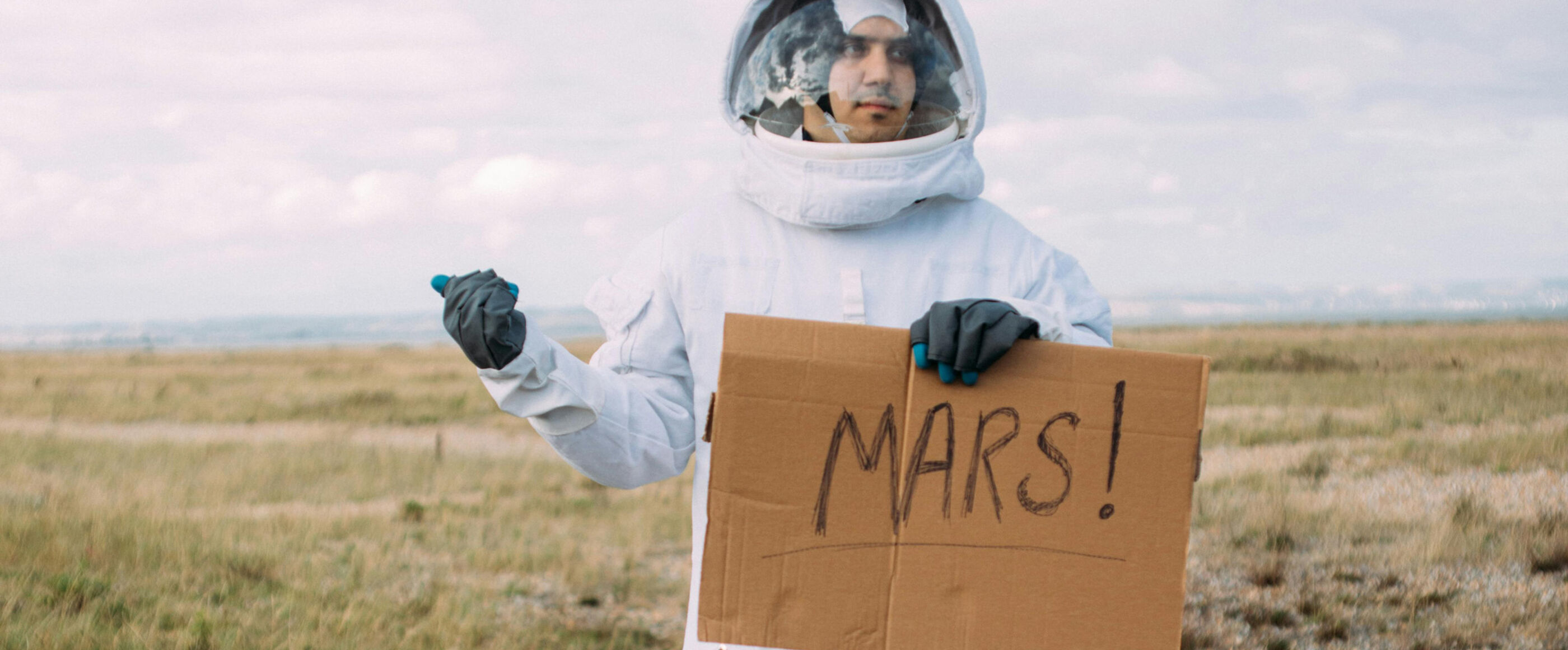 Eine als Mann lesbare Person in einem Raumanzug am Straßenrand und hält den Daumen raus. In der anderen Hand hält er ein Schild mit der Aufschrift MARS.