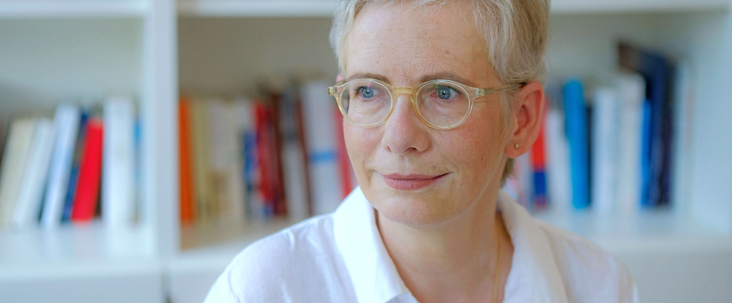 Der Bild zeigt eine weiblich lesbare Person mit kurzen hellblonden Haaren, einer hellen Brille, heller Hautfarbe und einer weißen Bluse. Die Person sitzt vor einem gefüllten Bücherregal.