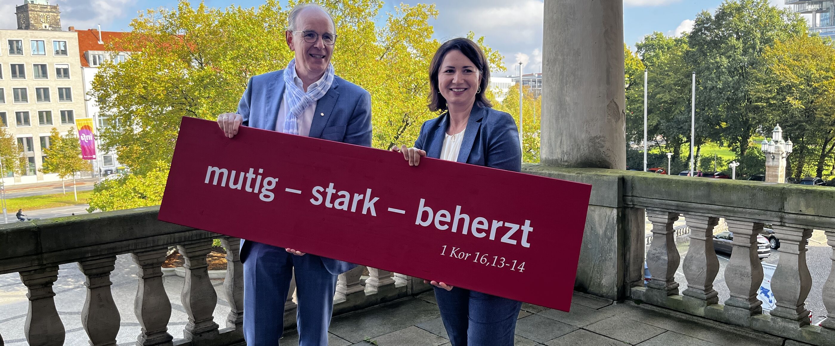 Auf einem steinernen Balkon stehen zwei Personen (eine männlich, ein weiblich gelesen) nebeneinander und halten gemeinsam eine rote Tafel in Händen. Darauf in weiß die Schrift: Mutig - stark - beherzt.