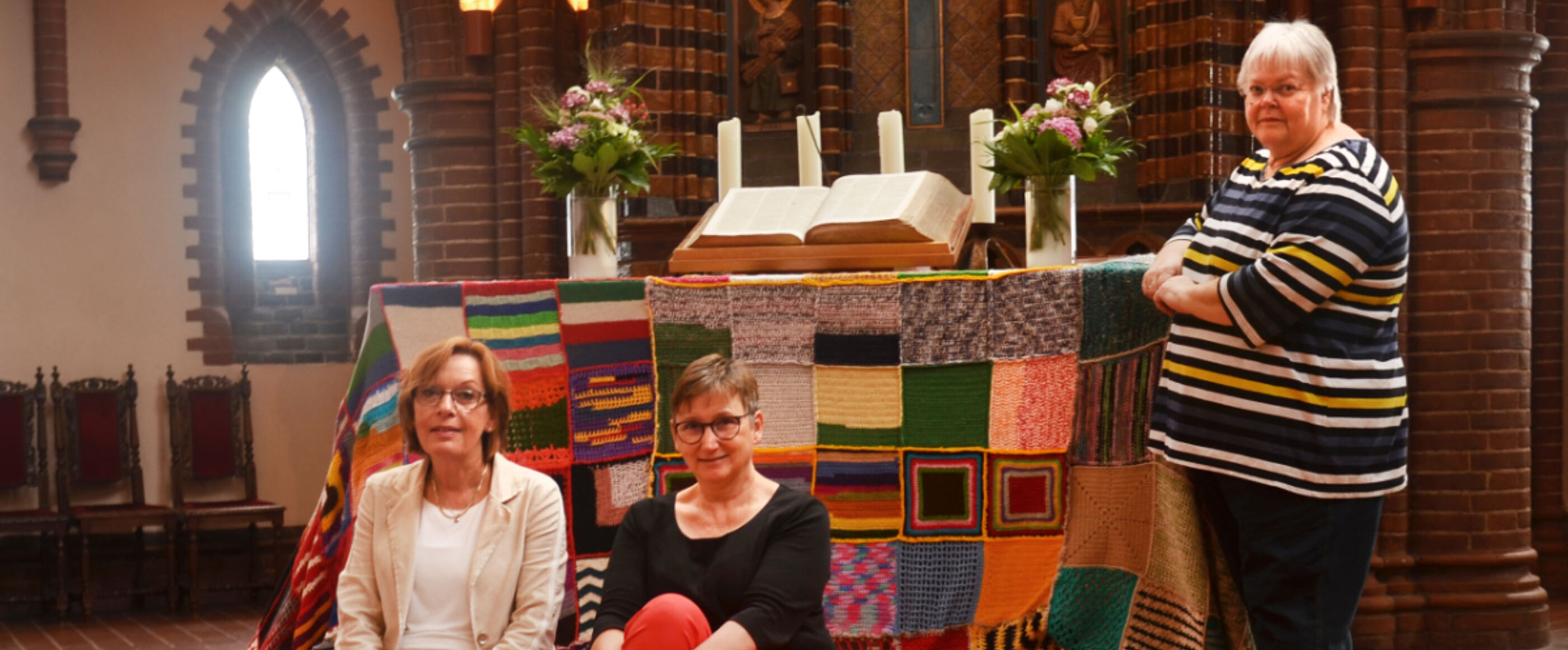 Zwei Frauen sitzen auf einer bunten Decke vor einem Kirchenaltar. Daneben steht eine weitere Frau.