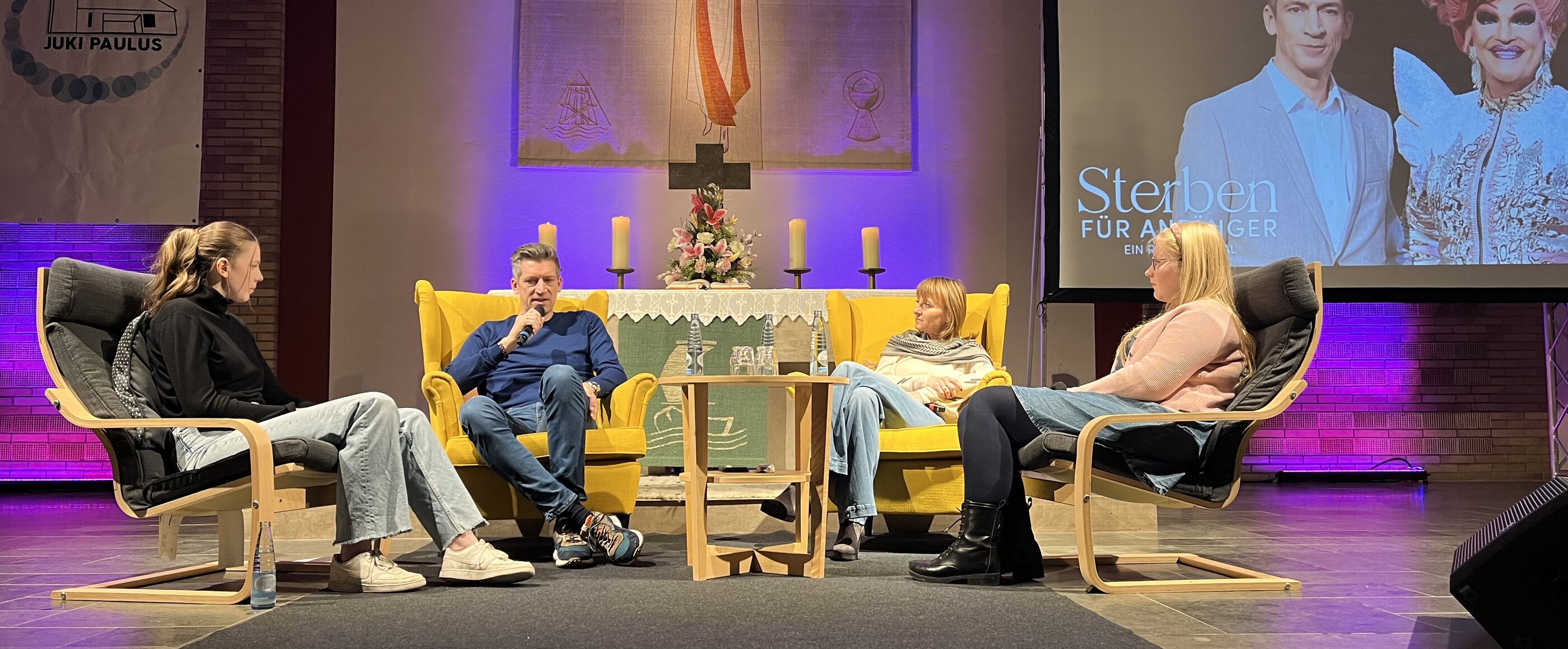 Vier Personen sitzen in Stühlen auf einer kleinen Bühne vor einem Altar. Rechts im Hintergrund ist eine Leinwand aufgebaut, auf der zwei Personen zu sehen sind: Steffen Hallaschka und Olivia Jones.