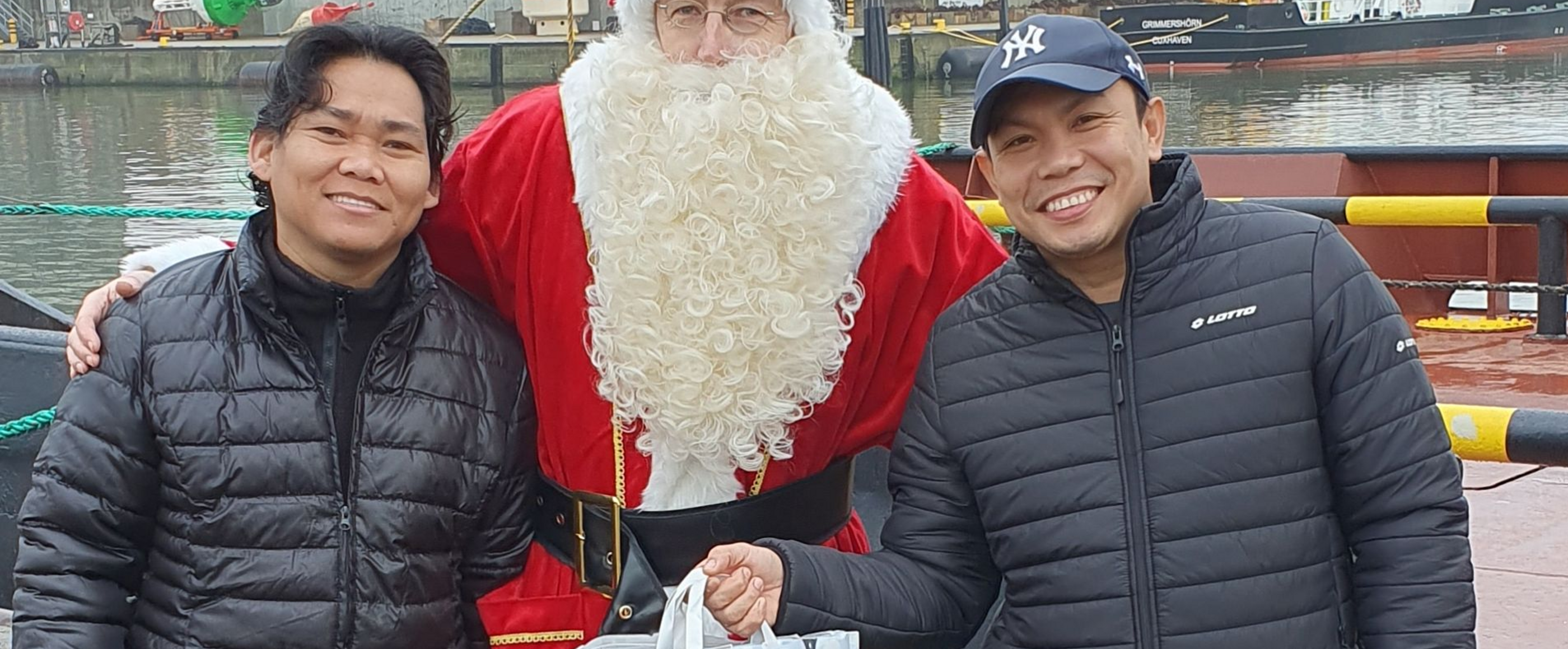 Zwei Männer mit Winterjacken halten Geschenktüten und stehen links und recht von einem Weihnachtsmann.