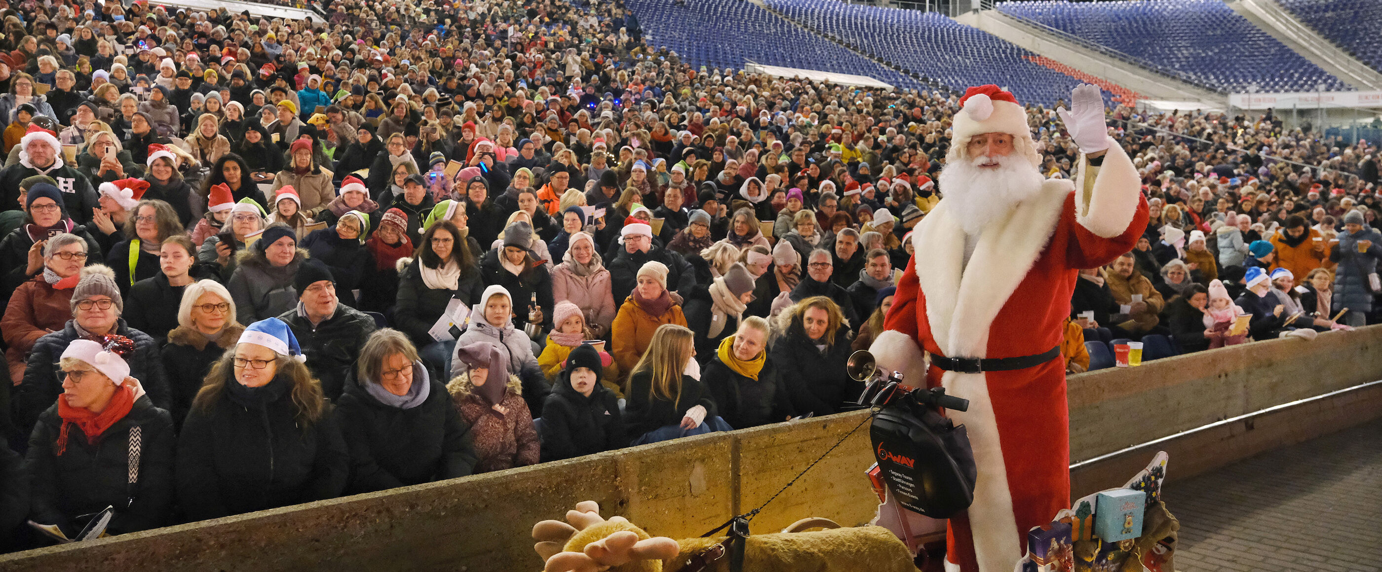 Ein Weihnachtsmann winkt vor einer Stadiontribüne voller Menschen.