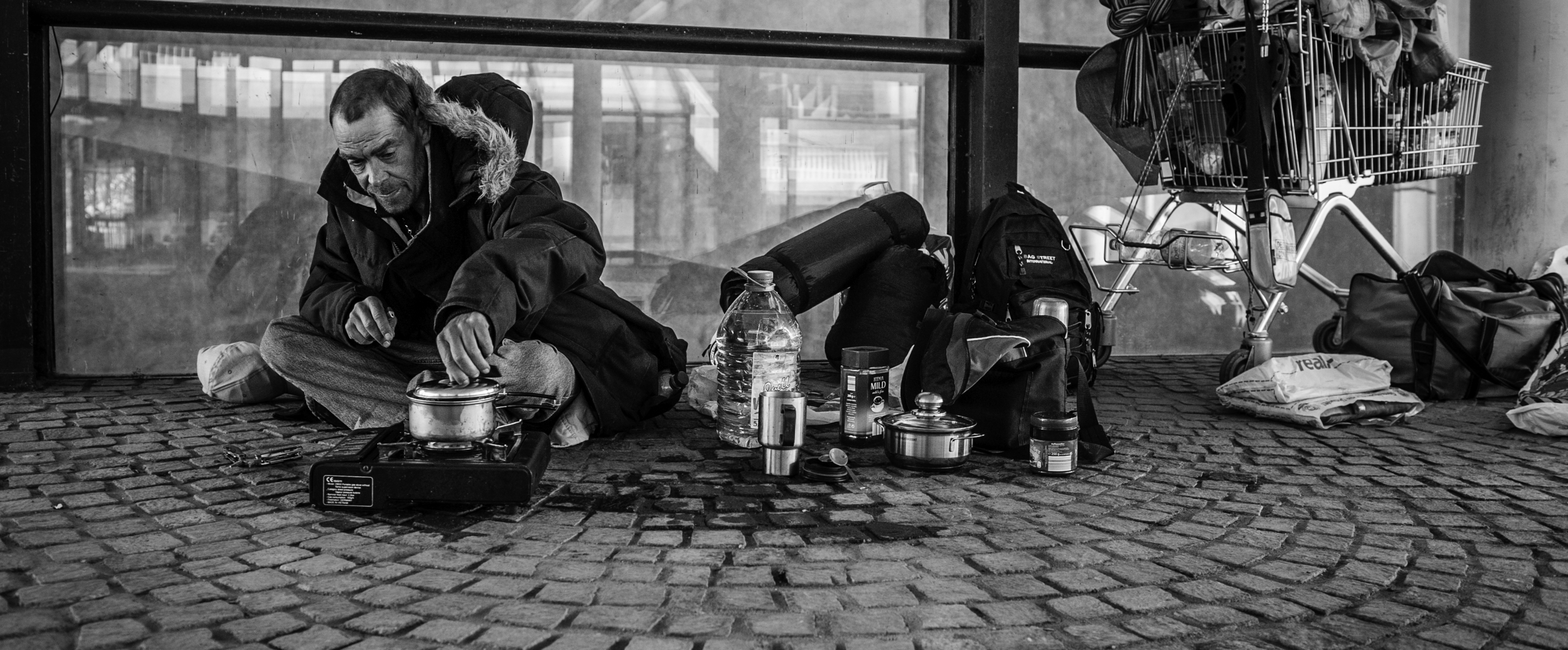 Ein schwarz-weiß-Bild zeigt einen wohl obdachlosen Mann, der mit einige Habseligkeiten auf der Straße vor einem Laden sitzt.