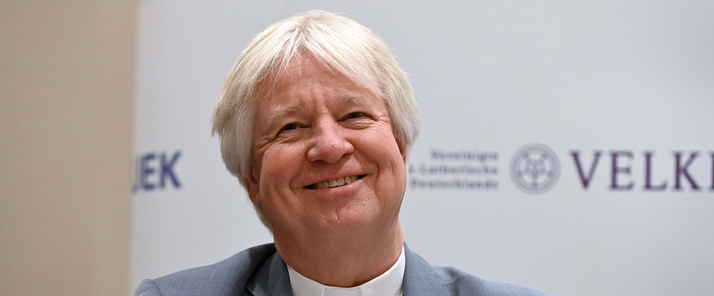 Ein Mann mit grauem Haar sitzt vor einer Stellwand mit der Aufschrift VELKD und UEK und lächelt.