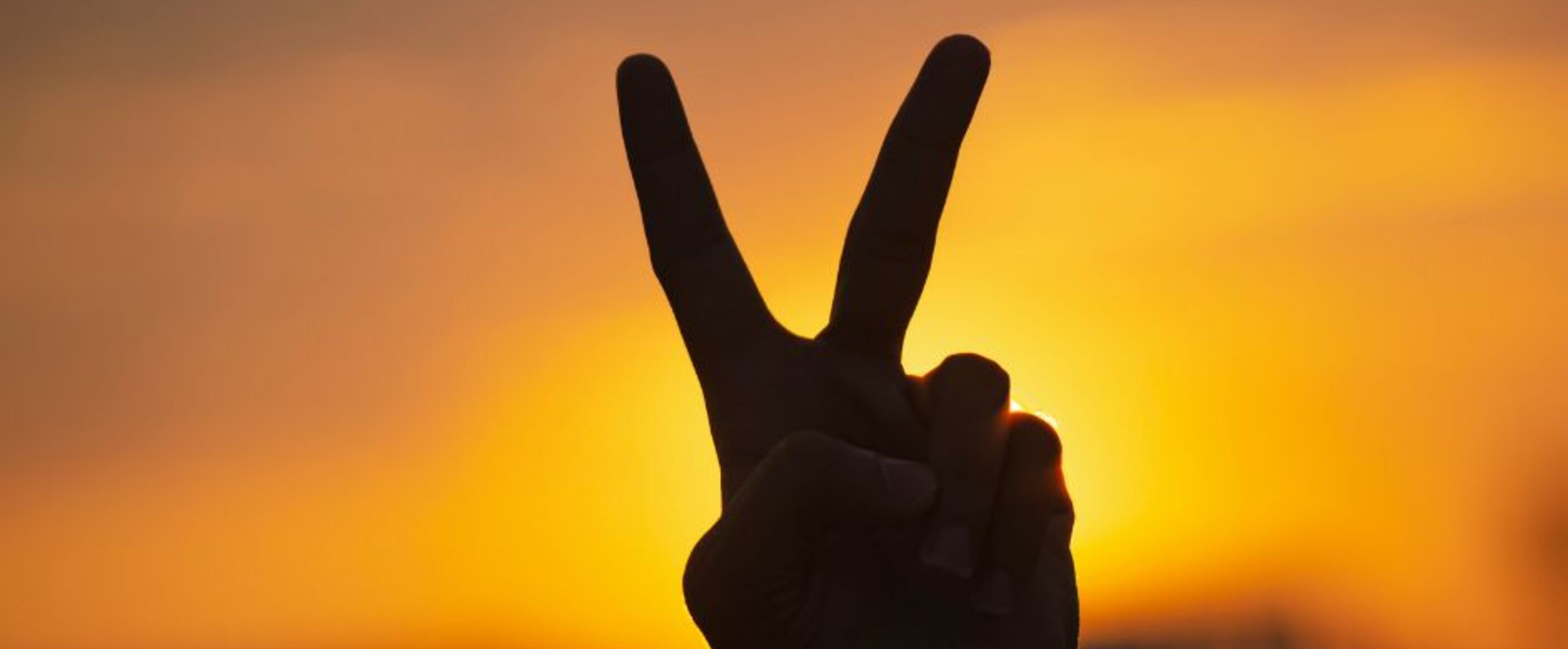 Eine Hand formt das Peace-Zeichen. Sie ist nur als Silhouette dunkel gegen einen orange leuchtenden Sonnenuntergang zu sehen.