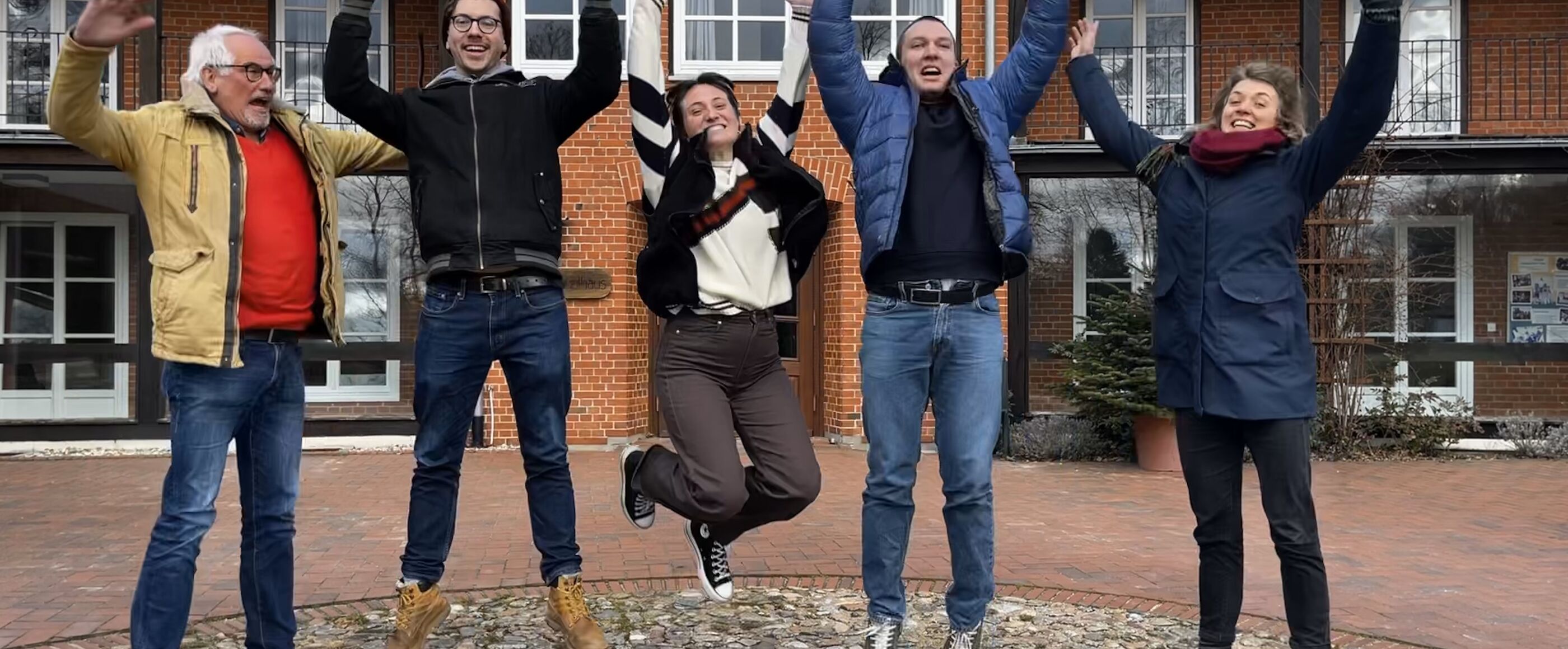 Fünf Menschen stehen auf einem Hofgelände im Freien und springen mit hochgestreckten Armen lachend in die Luft.