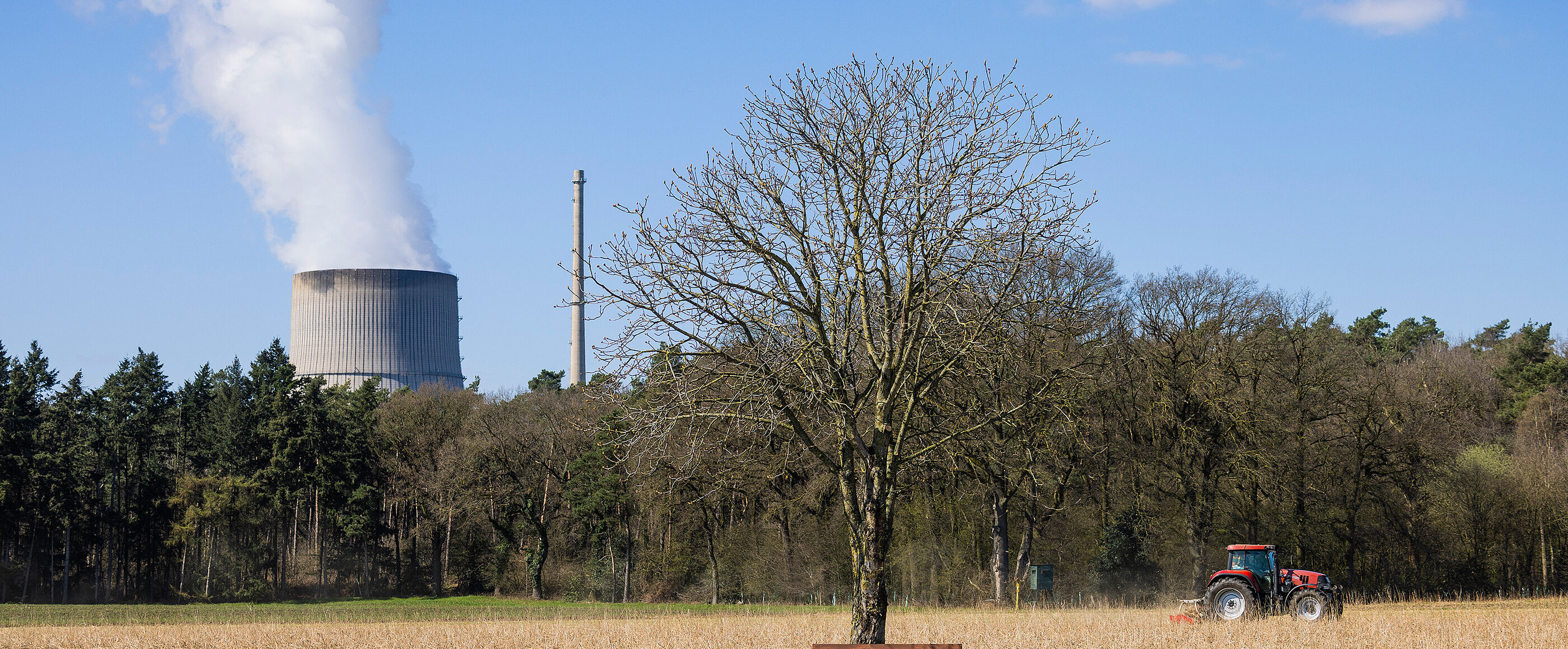 Eine Landschaft mit Baum und Bank; ein Trecker fährt über ein Feld, im Hintergrund quillt Dampf aus einem Kühlturm.