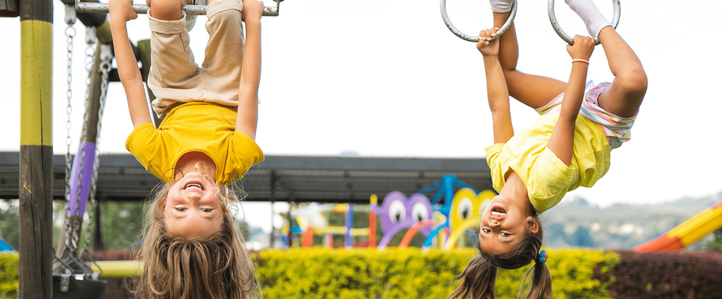 Zwei Mädchen mit gelben Shirts hängen kopfüber an Ringen und lachen breit.