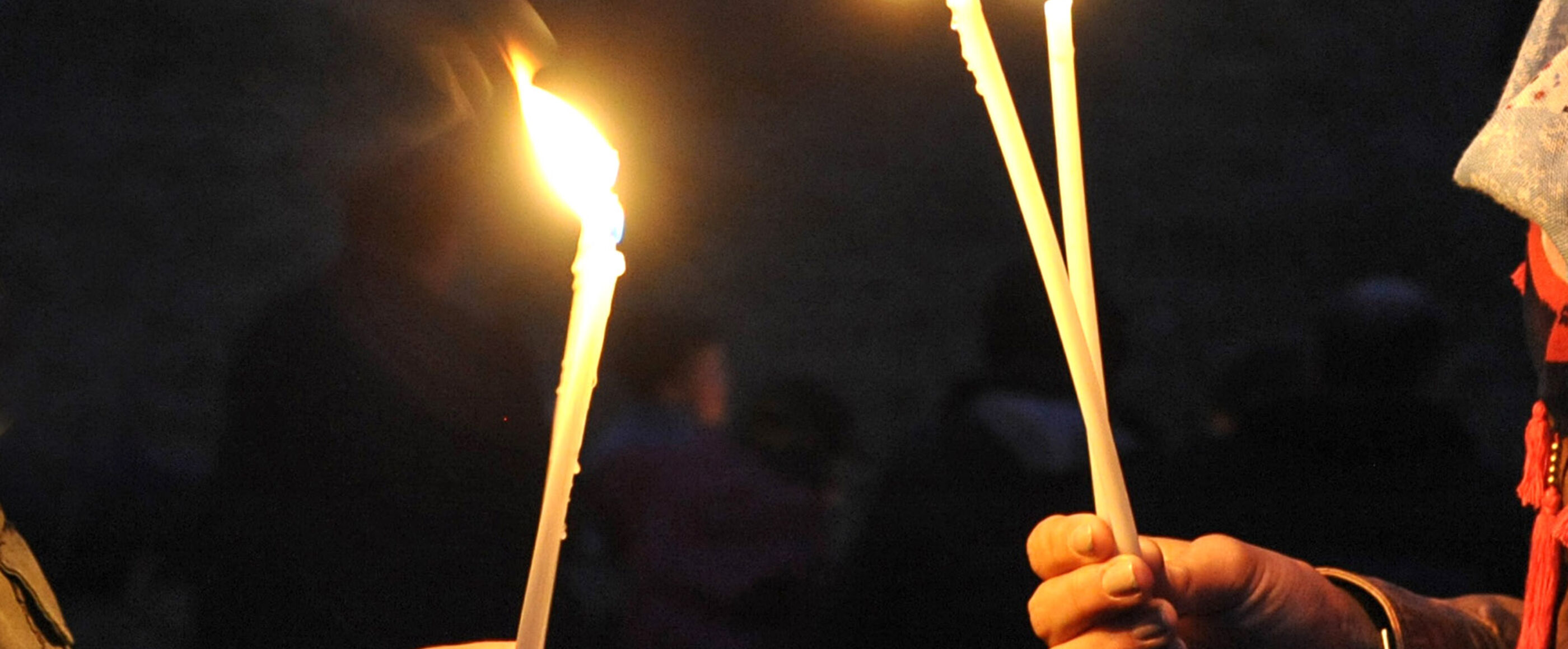 Zwei Menschen stehen sich gegenüber, zu sehen sind vor dunklem Hintergrund ihre Hände, die lange, dünne, brennende Kerzen halten.