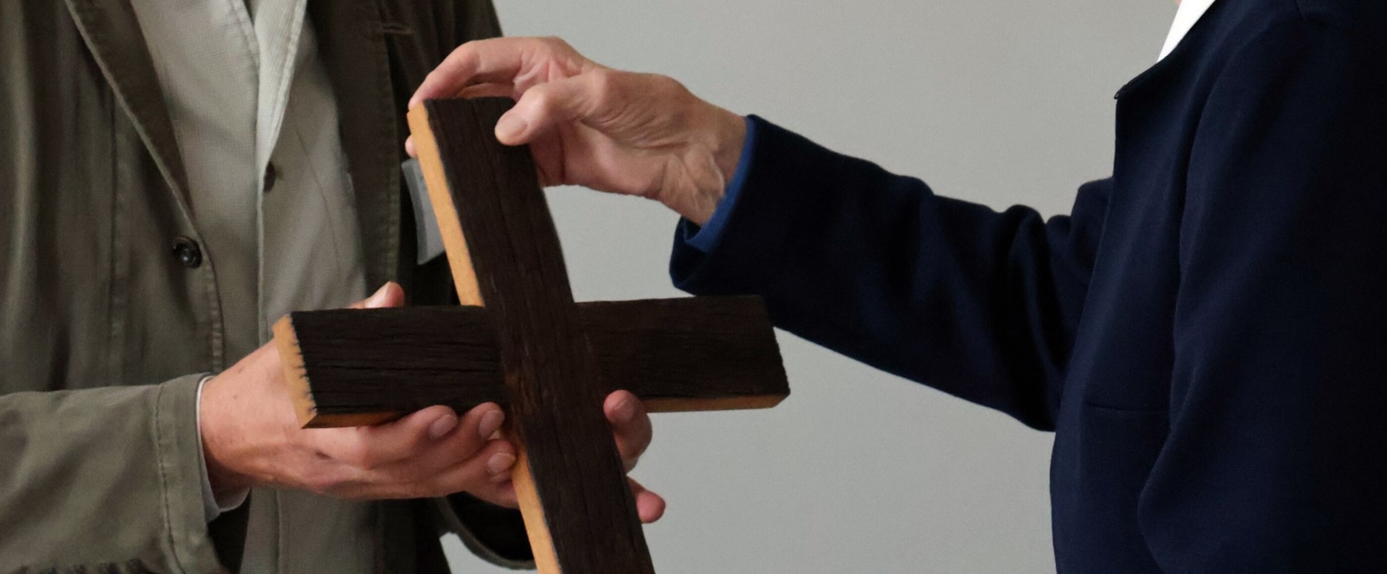 Eine Person übergibt einer anderen ein Holzkreuz. Sichtbar sind nur Hände und Schultern.
