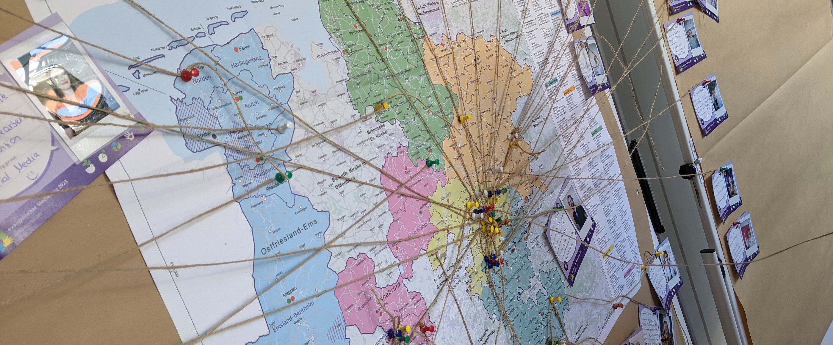 An einer braunen Pinnwand hängt eine bunte Karten von Niedersachsen. Bindfäden verbinden einige Orte mit lila Karten, auf denen Namen und Fotos sind. 