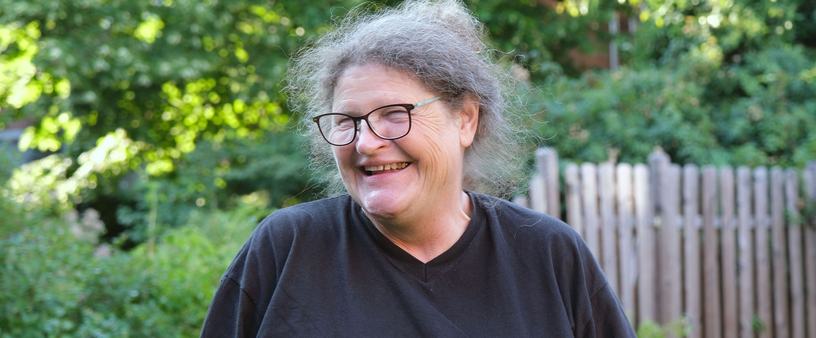 Eine Frau mit grauen Haaren, Brille und dunklem Shirt lacht, den Kopf nach rechts gedreht. Sie steht vor grünen Hecken und einem Holzzaun.