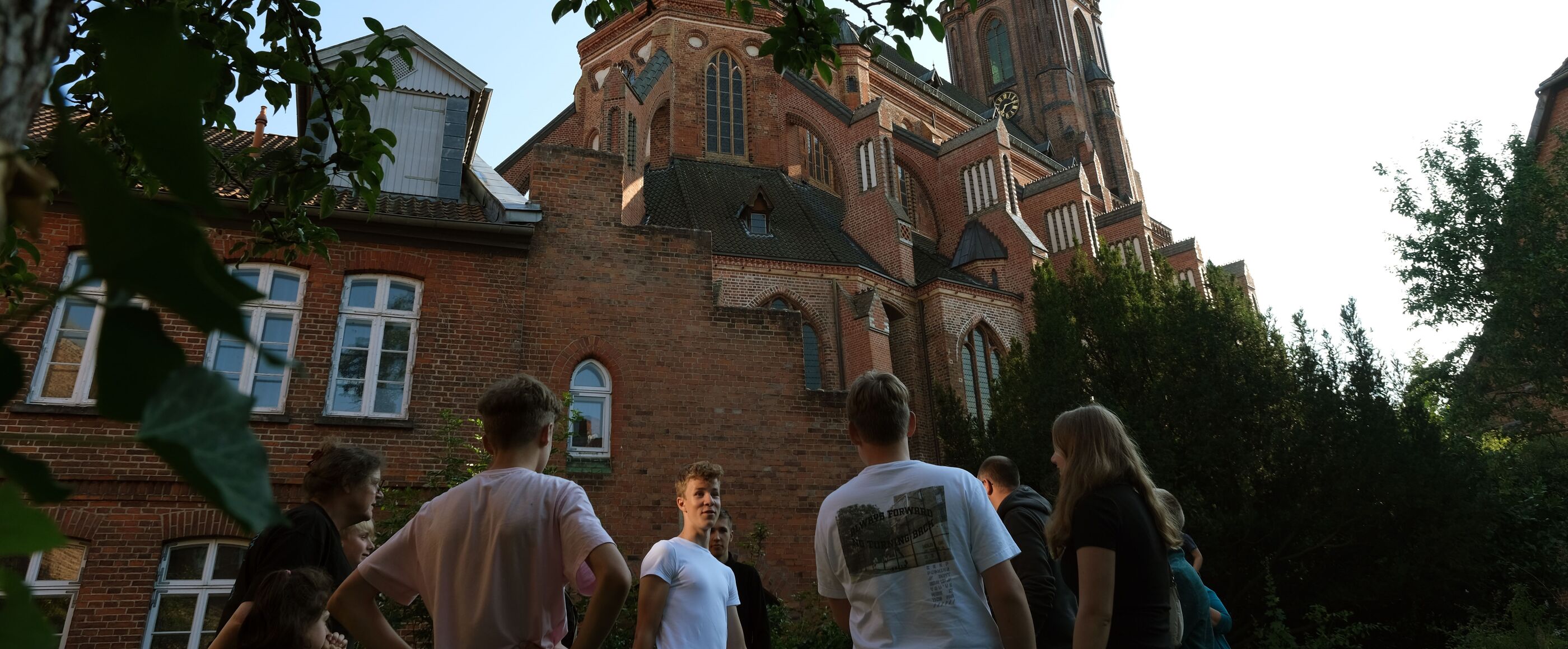 Mehrere junge Menschen stehen vor einer Kirche, sie sind von unten fotografiert. Die Sonne geht unter.