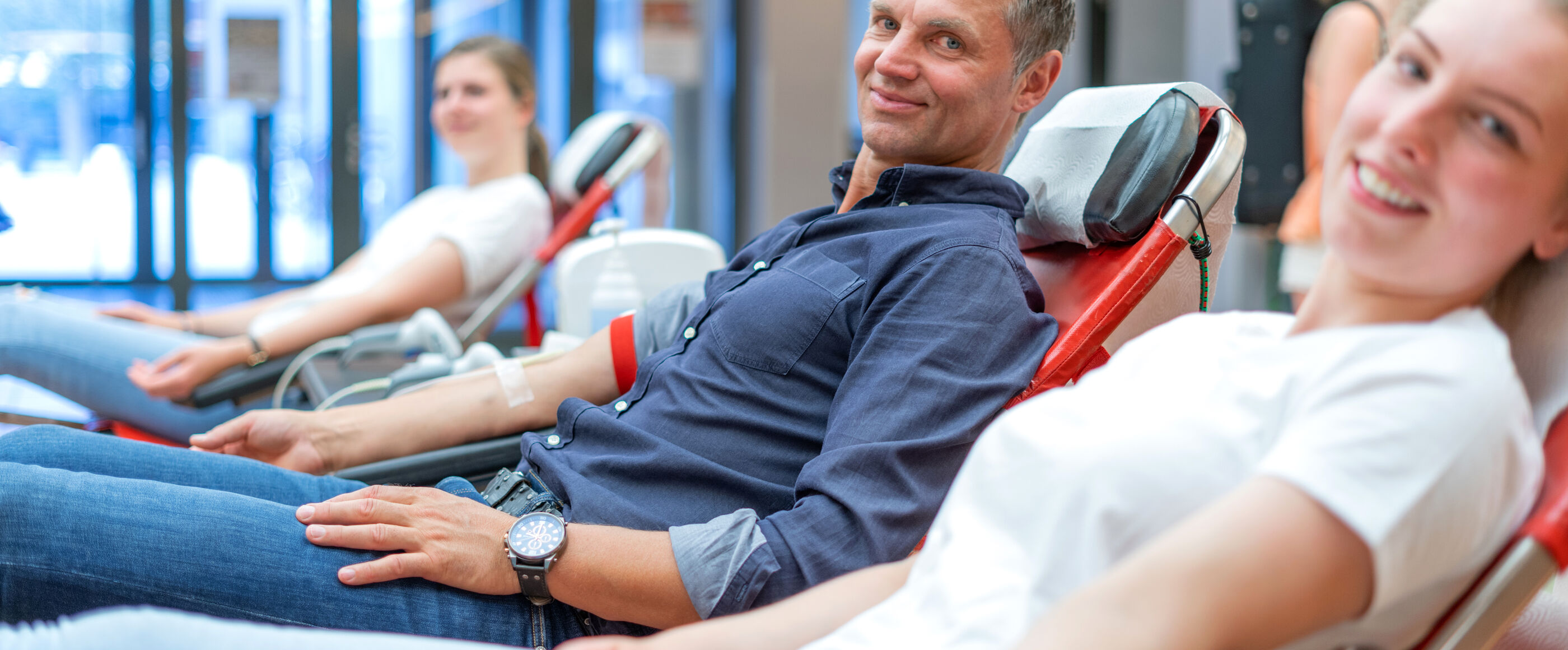 Drei Personen liegen nebeneinander und haben Schläuche in den Ellenbogen, zum Blutspenden. Sie lächeln. 
