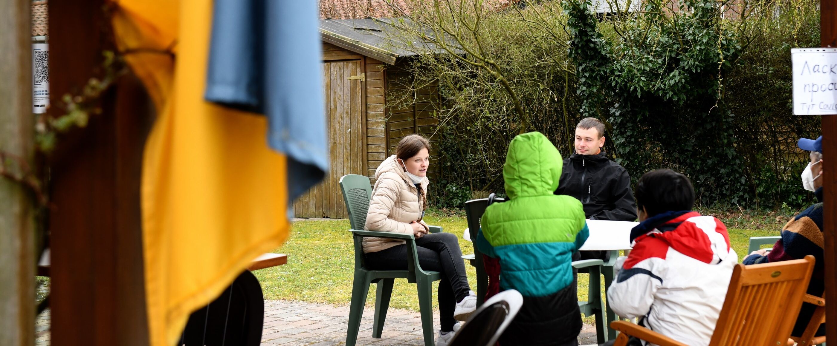 Fünf Menschen sitzen um einen Gartentisch auf einer Terrasse. Links, unscharf im Vordergrund, hängt eine blau-gelbe Fahne. 