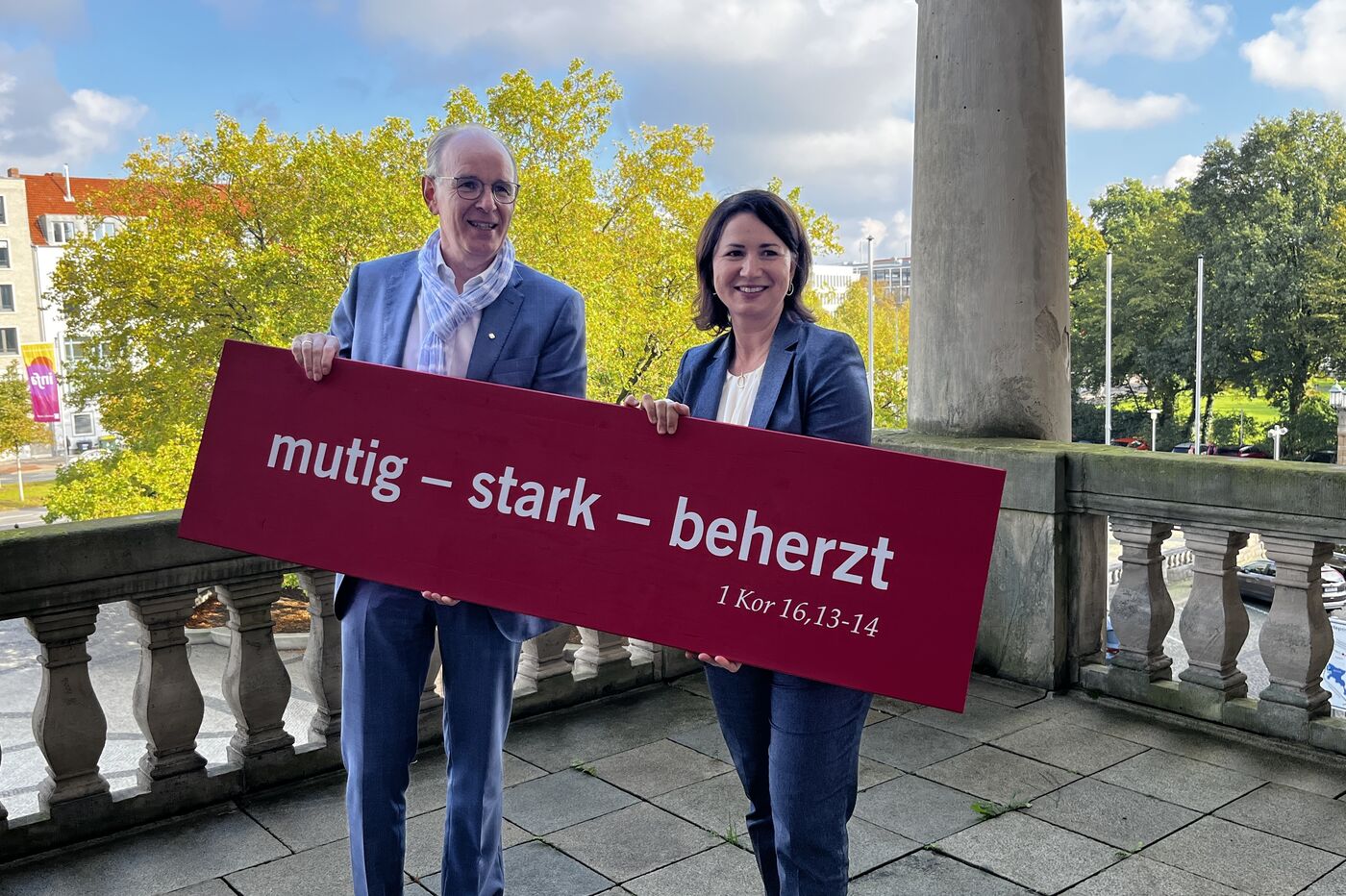 Auf einem steinernen Balkon stehen zwei Personen (eine männlich, ein weiblich gelesen) nebeneinander und halten gemeinsam eine rote Tafel in Händen. Darauf in weiß die Schrift: Mutig - stark - beherzt.