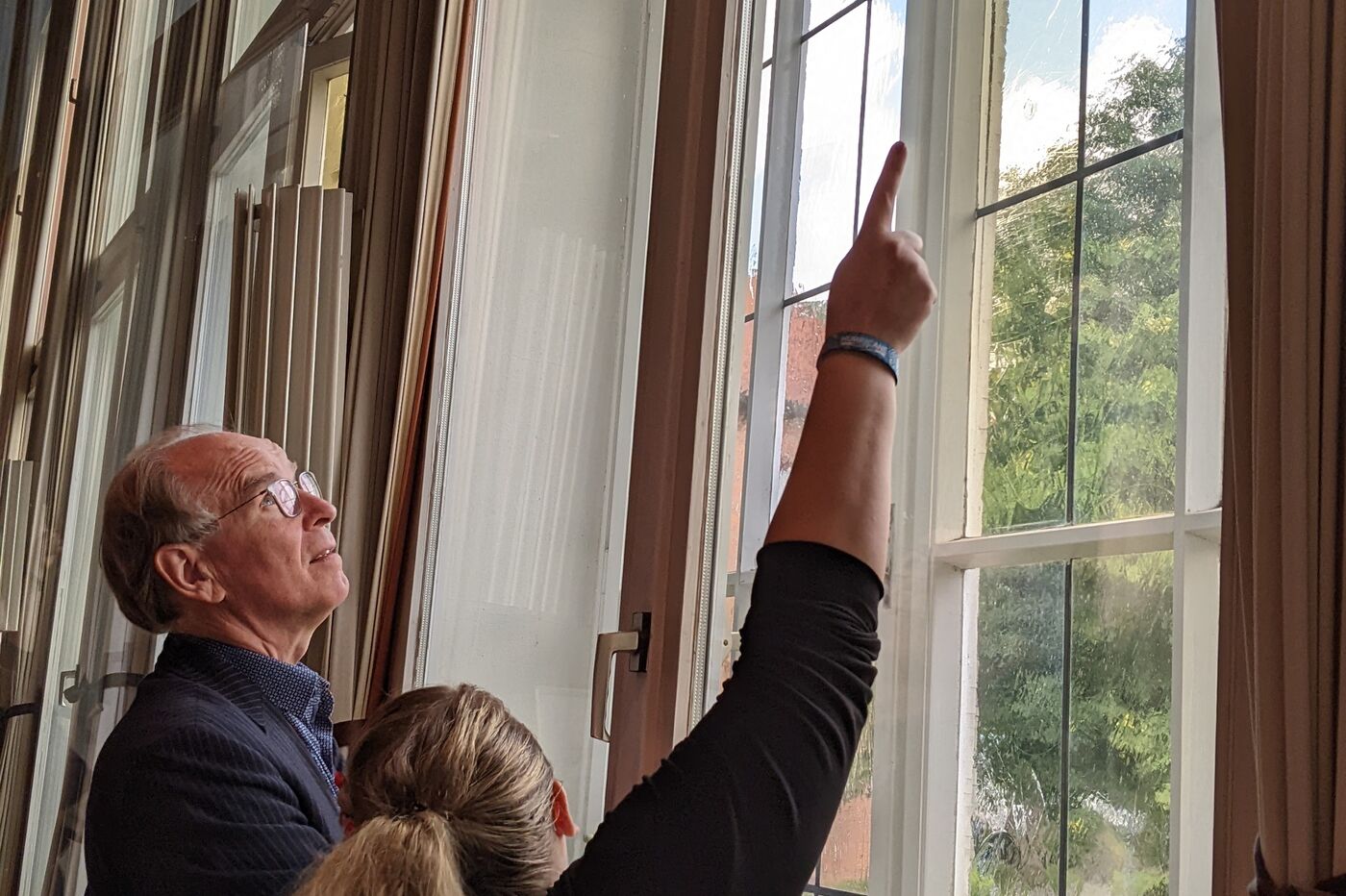 Ein älterer Mann und eine blonde Frau stehen an einem Fenster, die Frau zeigt auf etwas.