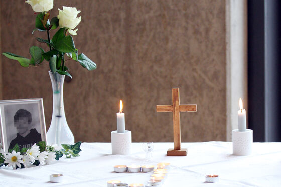 Das Bild zeigt einen Tisch mit einer weißen Tischdecke sowie darauf ein Schwarzweiß-Bild eines männlich lesbaren Kindes, eine Vase mit weißen Blumen, ein christliches Holzkreuz sowie zwei brennende Kerzen.