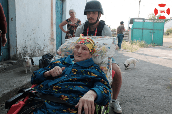 Das Bild zeigt eine ältere, weiblich lesbare Person in einem Rollstuhl, der von einer männlich lesbaren Person mit Militärhelm geschoben wird.