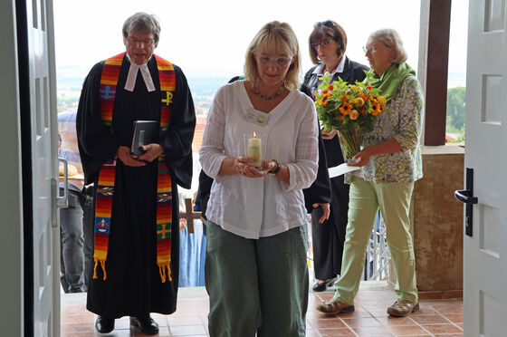 Vier Menschen betreten ein Haus: der Mann links trägt einen Talar und eine bunt gestreifte Stola, die Frau vorn in der Mitte eine weiße Bluse und trägt einen brennende Kerze in Händen. Recht folgen zwei Personen mit Blumenstrauß.