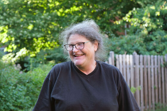 Eine Frau mit grauen Haaren, Brille und dunklem Shirt lacht, den Kopf nach rechts gedreht. Sie steht vor grünen Hecken und einem Holzzaun.