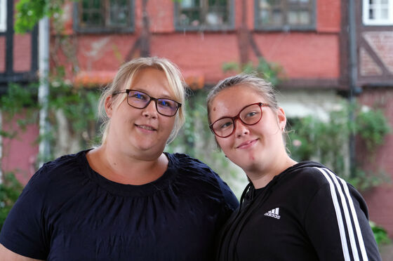 Zwei Frauen, die sich ähnlich sehen, Mutter und Tochter sind, lächeln zaghaft. beide tragen dunkle Oberteile.