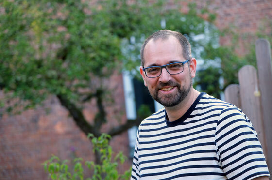 Ein Mann in gestreiftem Shirt mit Brille und Bart lächelt, im Hintergrund ein Baum vor einem Backsteinhaus.