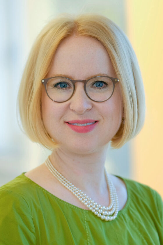 Das Bild zeigt eine weiblich lesbare Person mit blondem Pagenschnitt, brauner Brille, einer Perlenkette und einem grünen Oberteil.