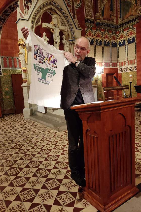 Ein Mann steht an einem Lesepult in einer Kirche und hält ein T-Shirt mit der Aufschrift „Peace“ hoch.