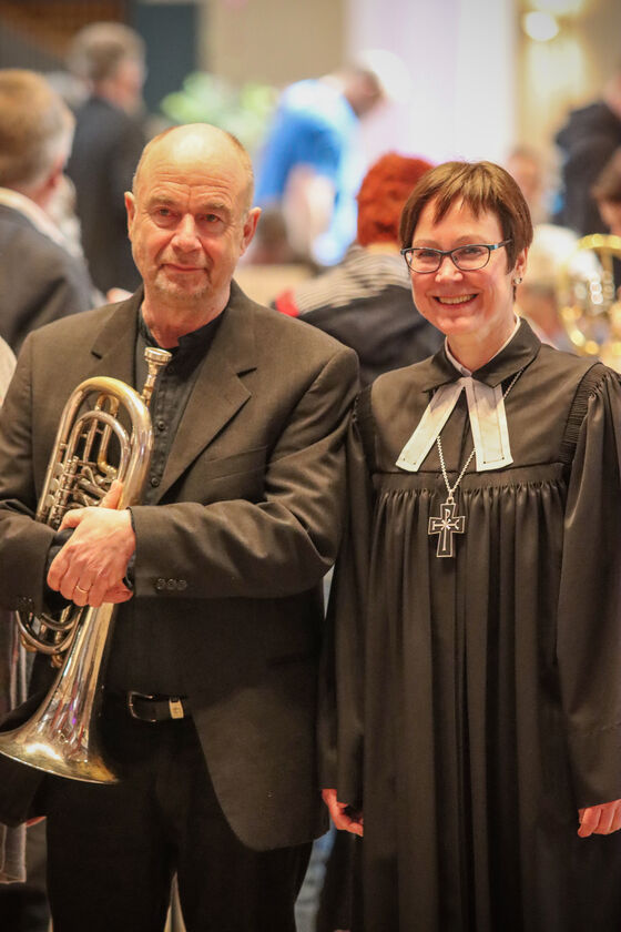 Eine männliche und eine weiblich gelesene Person stehen nebeneinander, der Mann hält eine Trompete in der Hand, die Frau trägt Talar.
