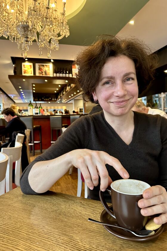 Eine Frau mit kurzen, braunen Haaren sitzt an einem Tisch in einem Café. Vor ihr steht eine braune Tasse mit Milchschaum.