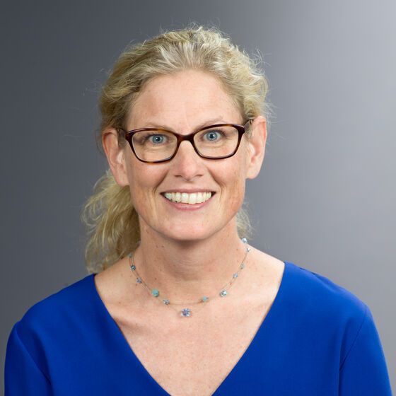 Eine blonde Frau, die Haare hinten zusammengenommen, mit Brille und blauem T-Shirt lächelt vor einem grauen Hintergrund.