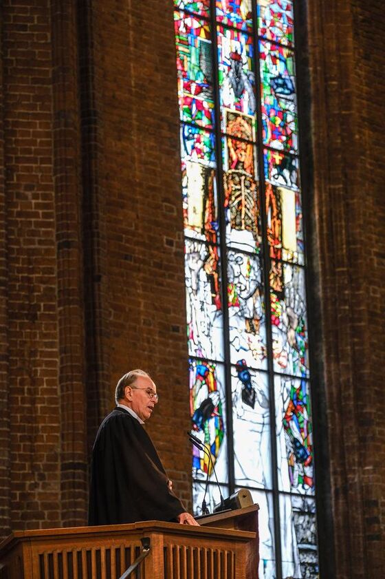 Eine männlich lesbare Person im Talar auf einer Kanzel, im Hintergrund ein großes buntes Kirchenfenster, das Martin Luther zeigt.