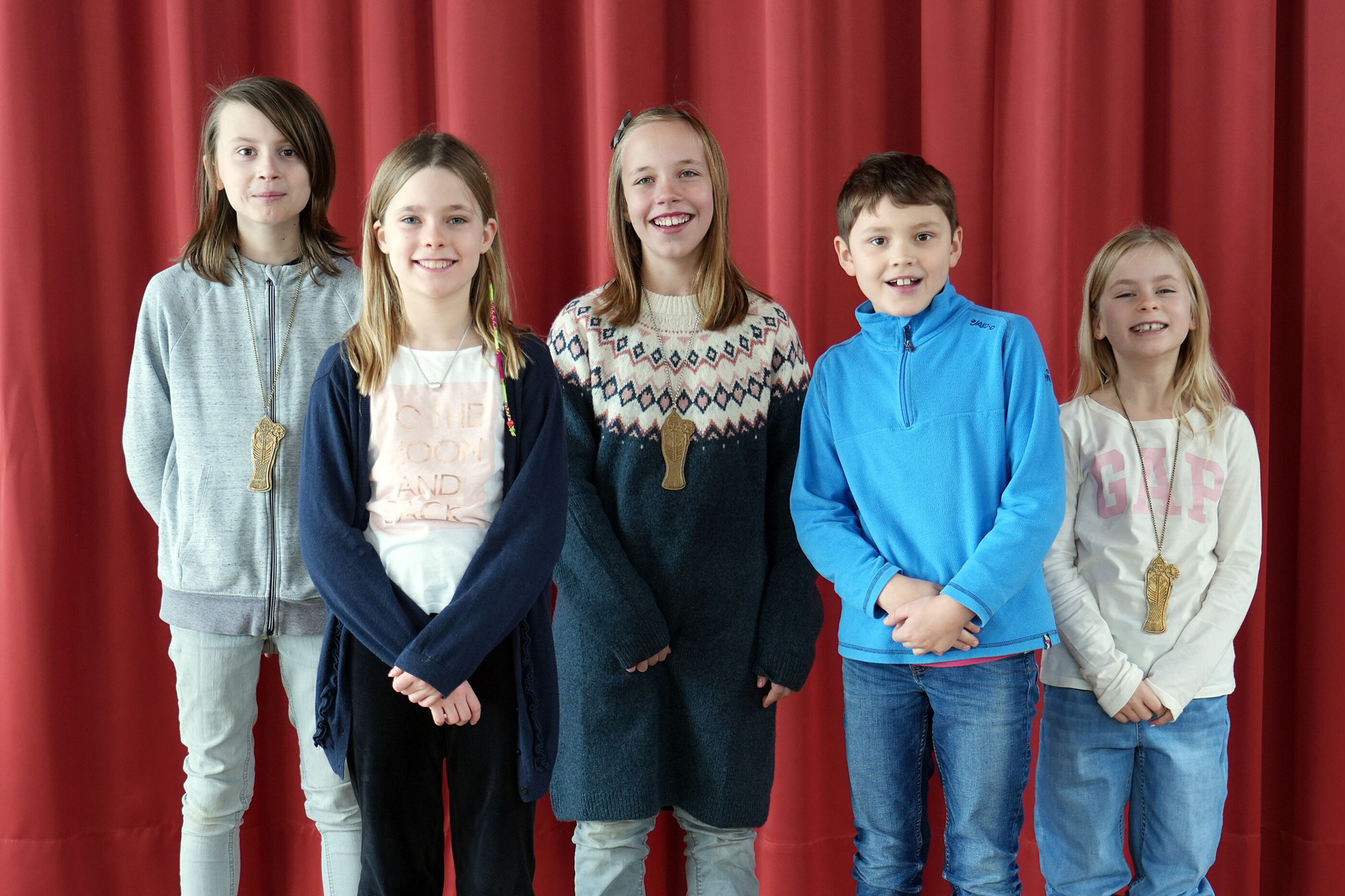 Fünf Kinder stehen vor einem roten Vorhang und lächeln in die Kamera.