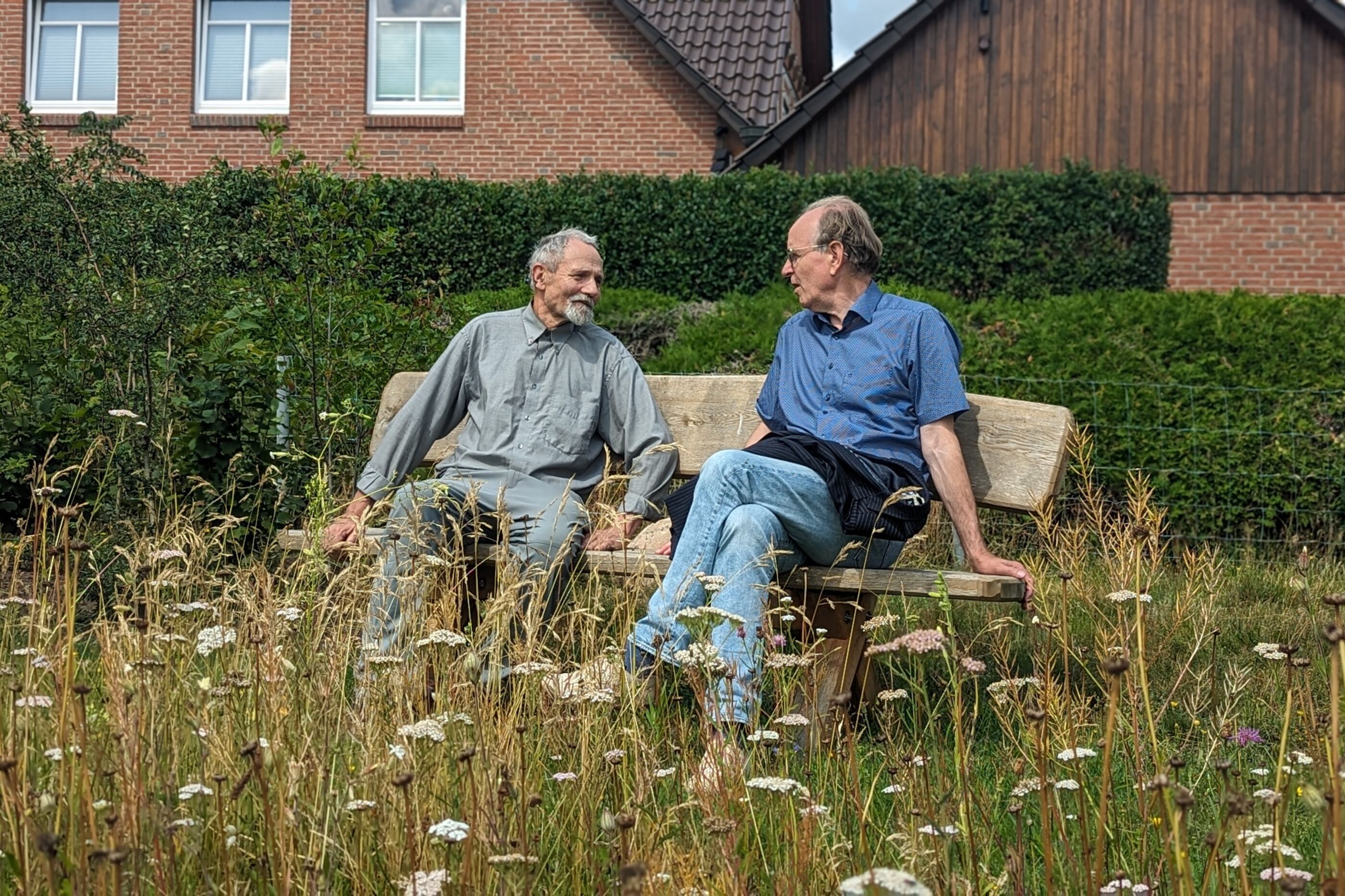 Zwei ältere Männer sitzen auf einer Bank, im Vordergrund blühende Gräser. Im Hintergrund ein Backstein-Einfamilienhaus.