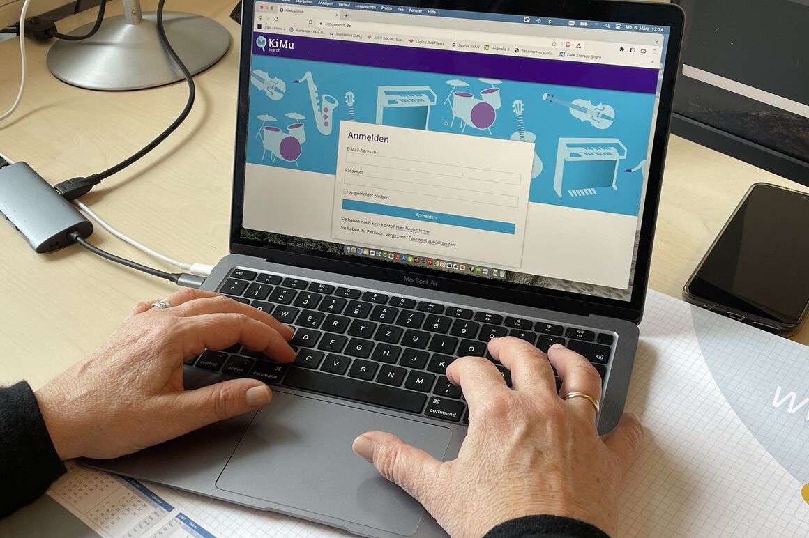 Zwei Hände schreiben auf einer Laptop-Tastatur. Auf dem Bildschirm ist die Startseite des Portals KiMuSearch zu sehen.