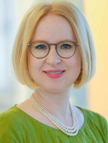 Das Bild zeigt eine weiblich lesbare Person mit blondem Pagenschnitt, brauner Brille, einer Perlenkette und einem grünen Oberteil.