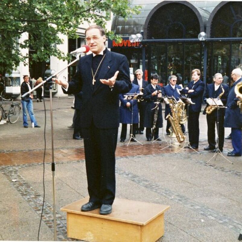 Das Bild zeigt eine männlich lesbare Person in schwarzem Anzug, die auf einem Platz auf einem kleinen Podest stehend in ein Mikrophon spricht.