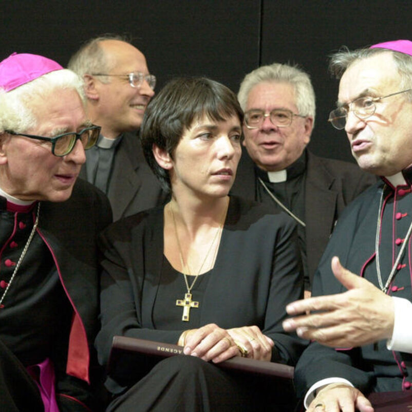 Das Bild zeigt eine weiblich lesbare Person mit kurzen dunklen Haaren dicht bedrängt von einer Gruppe katholischer Geistlicher, die um sie herum sitzen und mit ihr sprechen.