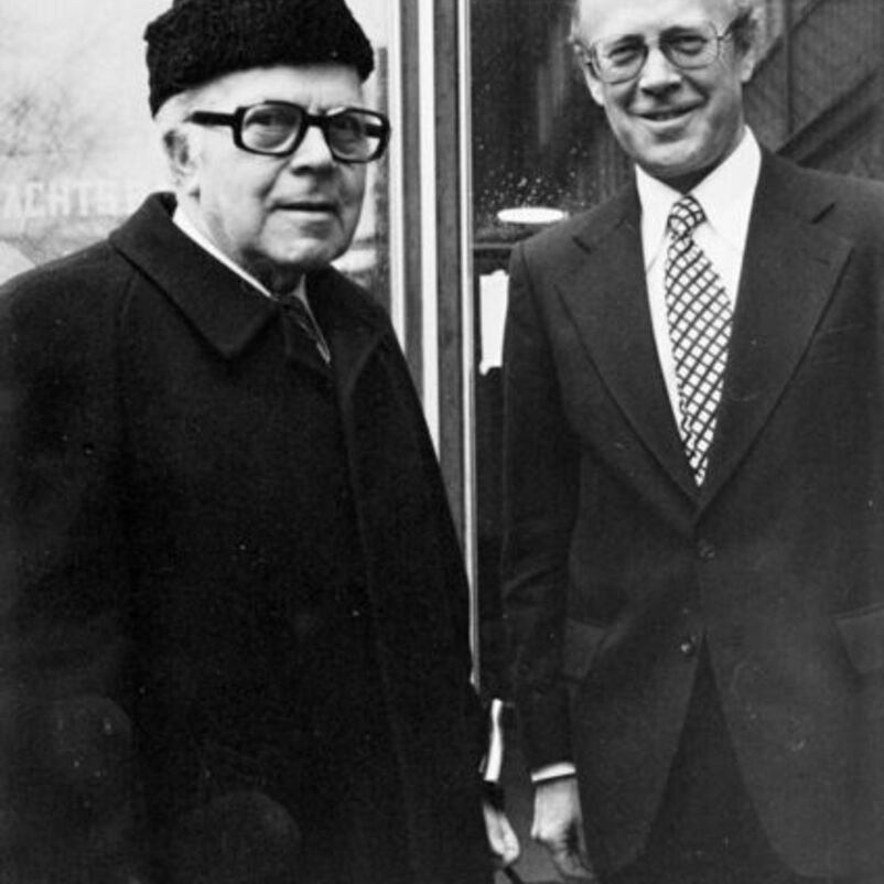 Das Bild zeigt zwei männlich lesbare Personen, die linke im Bild hat einen schwarzen Mantel und eine schwarze Kappe auf dem Kopf, die rechte trägt einen Anzug mit Krawatte und weißem Hemd.