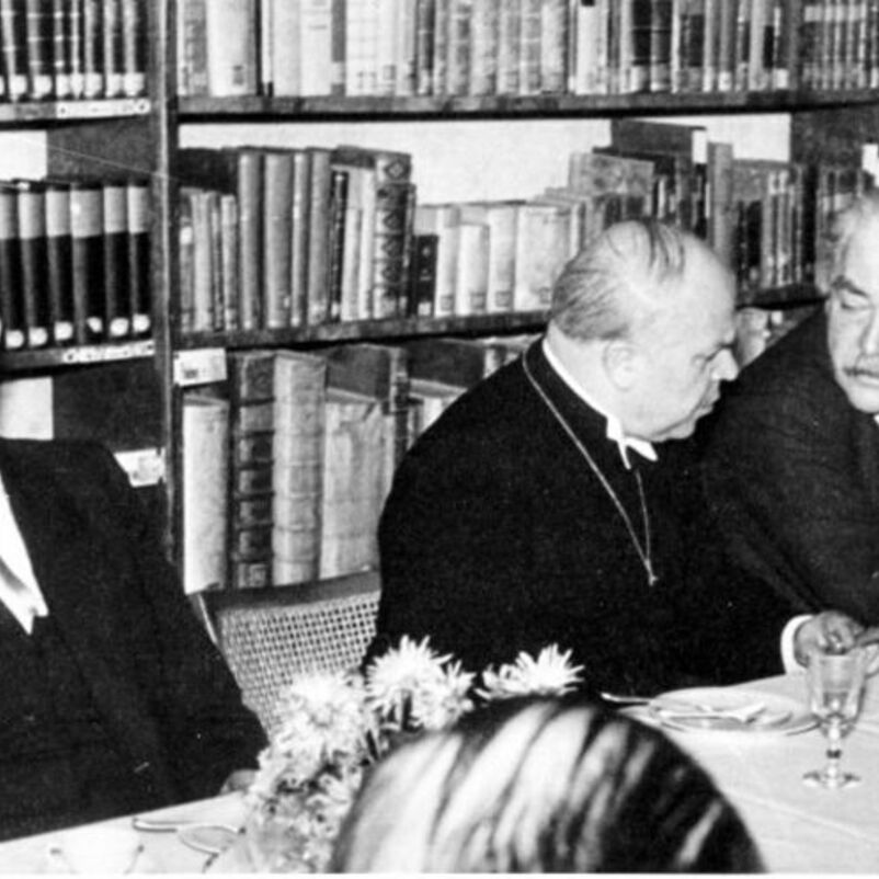 Das Schwarzweiß-Bild zeigt drei männlich lesbare Personen nebeneinander an einem Tisch, auf dem Gläser stehen. Im Hintergrund ist ein dicht gefülltes Bücherregal. 