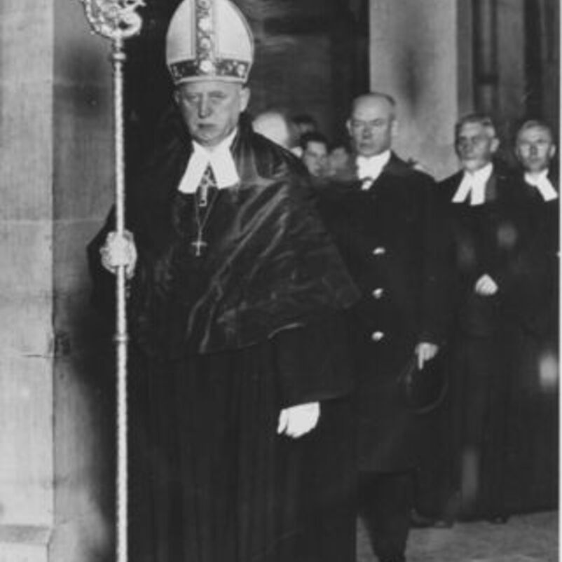 Das Schwarzweiß-Bild zeigt eine männlich lesbare Person mit Bischofsstab laufend durch einen Kirchgang vor einer Gruppe ihm folgender Menschen in schwarzen Anzügen.