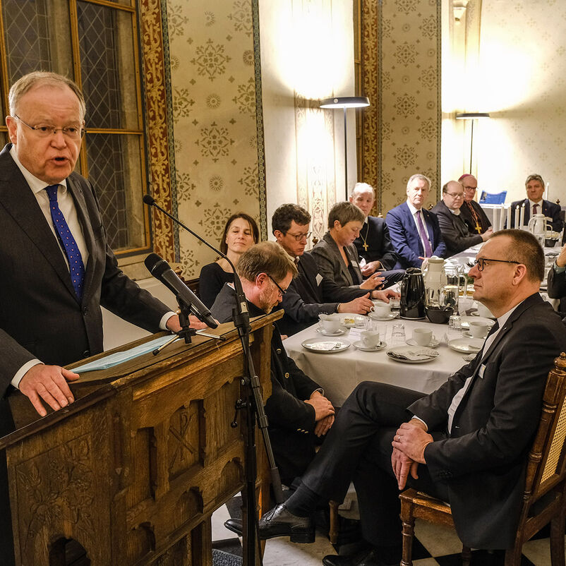 Ein Mann im Anzug mit blauer Krawatte steht an einem Rednerpult. Links von ihm sitzen etwa ein Dutzend Menschen an einem gedeckten Tisch.