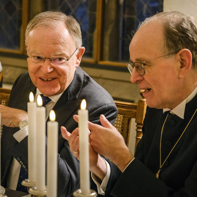 Zwei Männer sitzen nebeneinander und gestikulieren. Vor ihnen ein gedeckter Tisch mit Kerzenleuchtern.