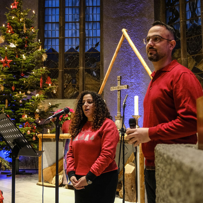 Eine schwarze Frau und ein Mann stehen an Mikrophonen vor einem großen Weihnachtsbaum in einer Kirche.