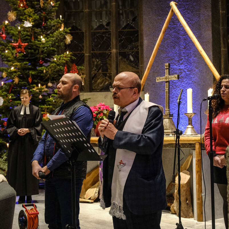 Vier Personen stehen an Mikrophonen, einer liest von einem Notenständer. Im Hintergrund stehen ein großer Weihnachtsbaum und ein Kreuz.
